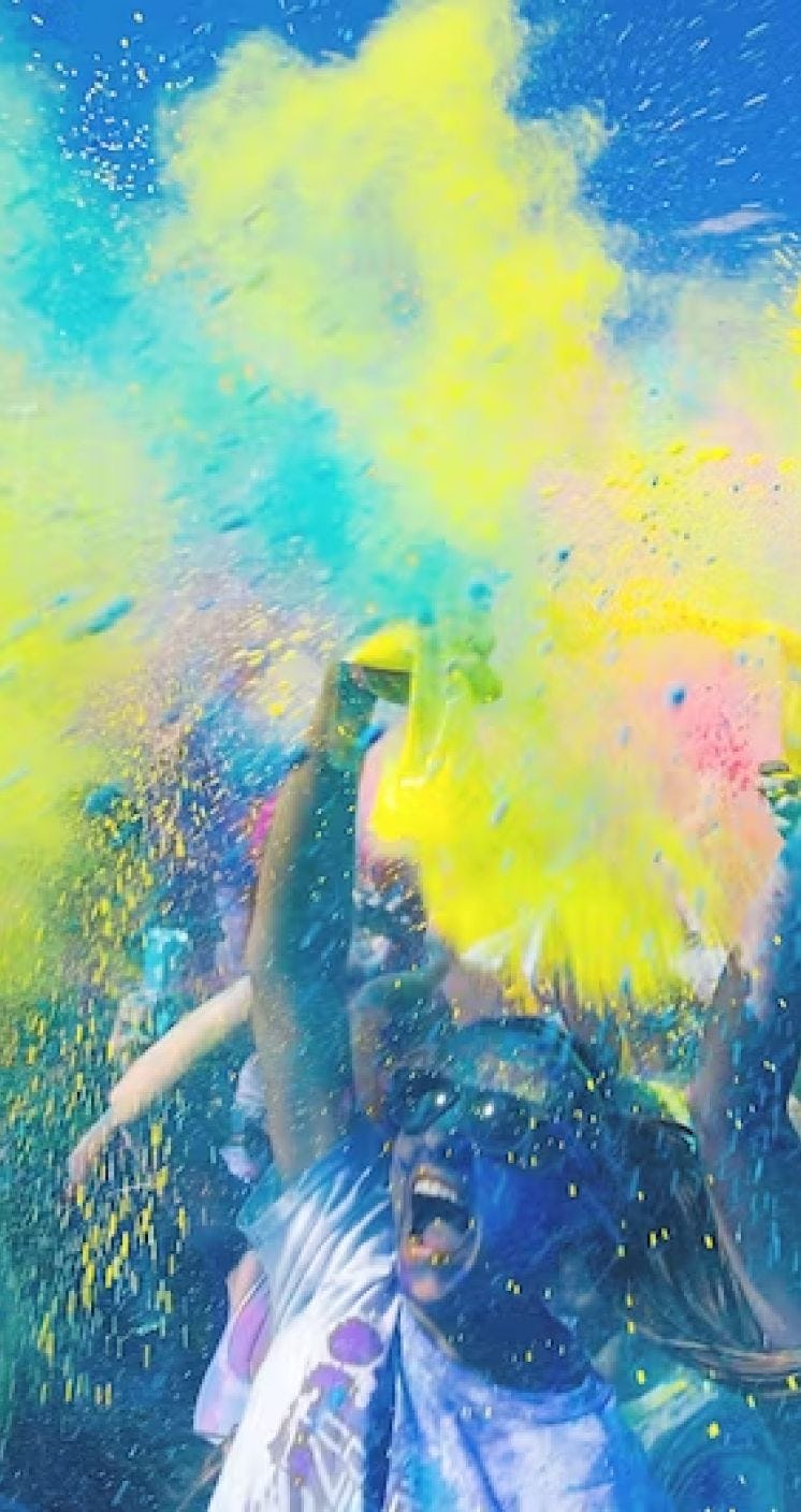 Large group of people covered in colorful powders with clouds of teal and yellow and pink powder filling the air and covering their bodies.