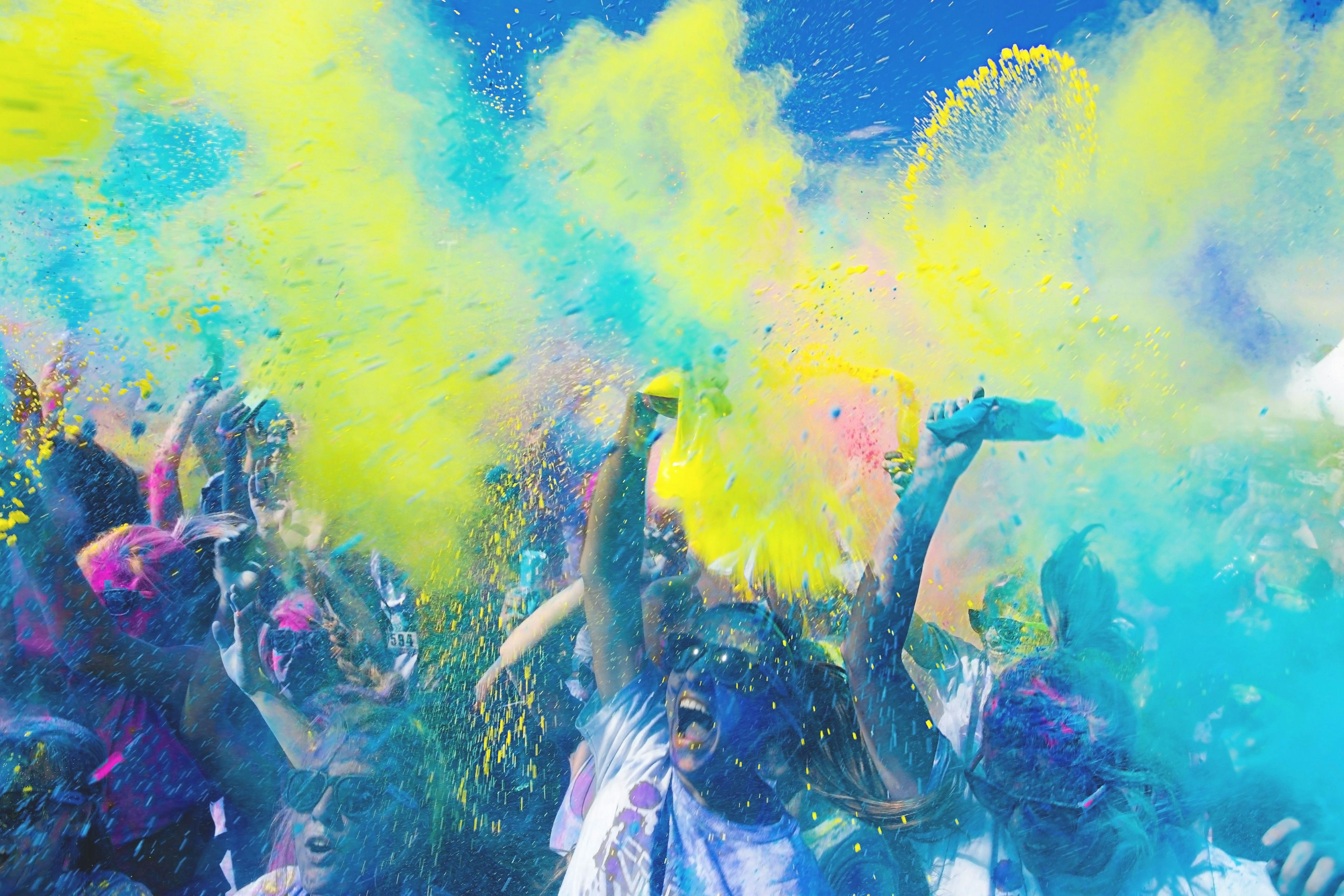 Large group of people covered in colorful powders with clouds of teal and yellow and pink powder filling the air and covering their bodies.